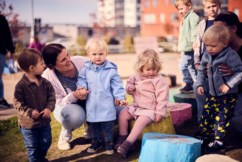 Förskolebarn sitter på gården med två pedagoger.